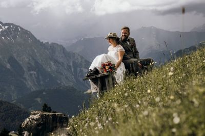 Brautpaar sitzt auf einer Holzbank in den Bergen, umgeben von Wiesenblumen und Alpenpanorama.