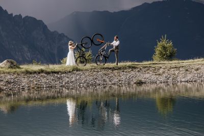 Braut und Bräutigam mit Fahrrädern am See, die Reifen aneinanderstoßend – kreative Hochzeitsaufnahme.