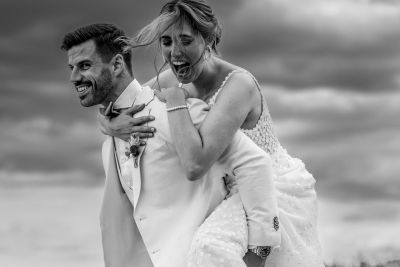 Bräutigam trägt seine lachende Braut auf dem Rücken – humorvolles Schwarz-Weiß-Hochzeitsfoto.