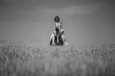 Schwarz-weiß-Foto eines Paares auf einem Feld – die Braut sitzt auf den Schultern des Bräutigams, verspielter Moment.