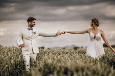 Brautpaar geht durch ein Getreidefeld, hält sich an den Händen und lacht – emotionale Sommerhochzeit.
