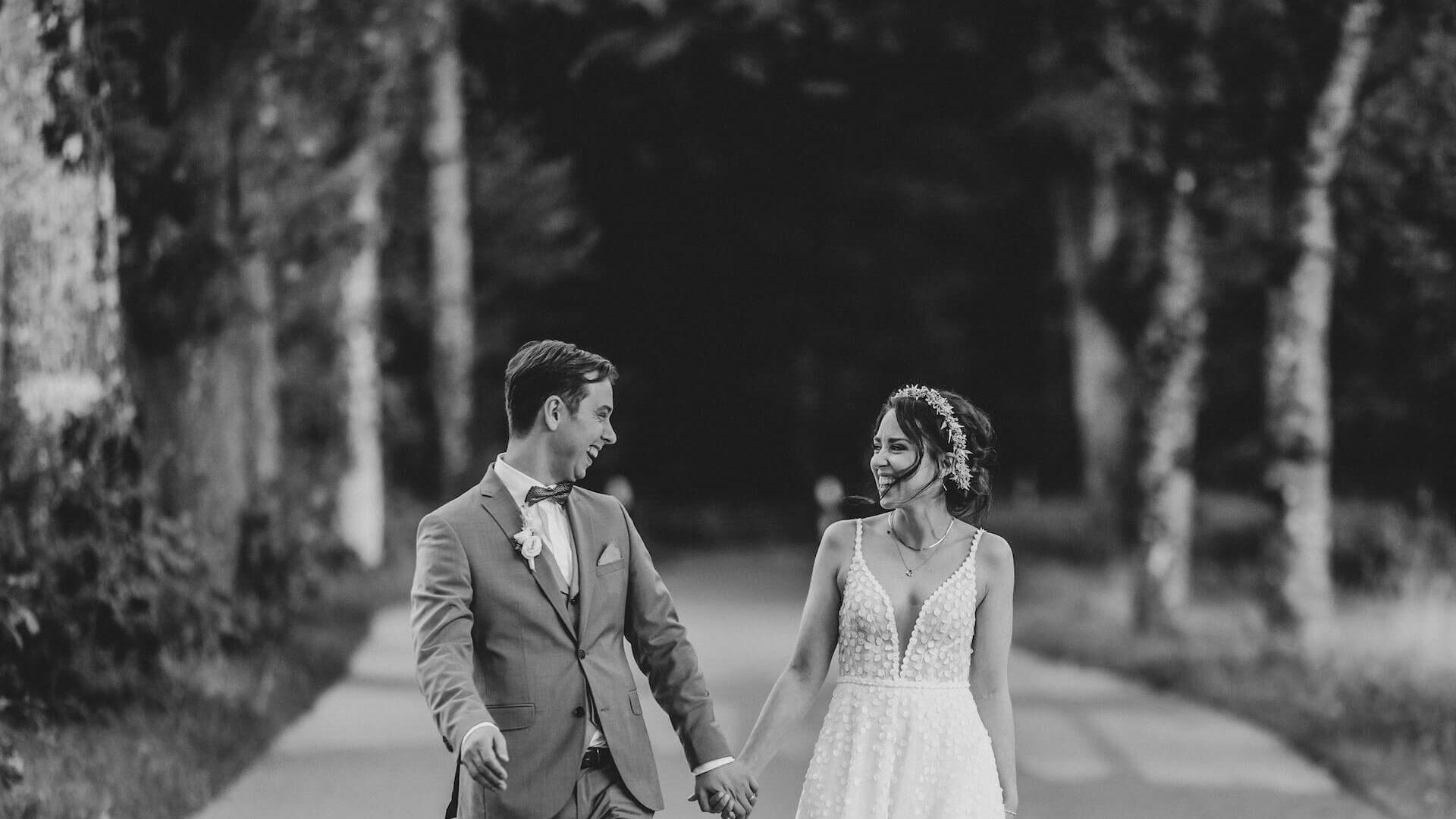 Brautpaar geht lachend Hand in Hand auf einer Allee – romantisches Schwarz-Weiß-Foto.