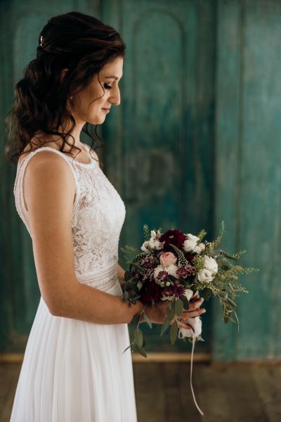 Braut steht mit Blumenstrauß vor türkisfarbener Wand – klassisches Porträt in natürlichem Licht.