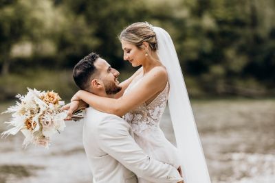 Bräutigam hebt Braut in den Armen, beide lächeln sich liebevoll an – romantisches Hochzeitsfoto.