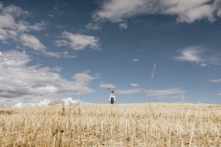 Braut wir über ein weites, goldenes Feld getragen und hält den Blumenstrauß freudig in die Luft – Hochzeitsfoto mit weitem Himmel und natürlicher Leichtigkeit.