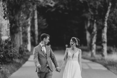 Brautpaar geht lachend Hand in Hand auf einer Allee – romantisches Schwarz-Weiß-Foto.
