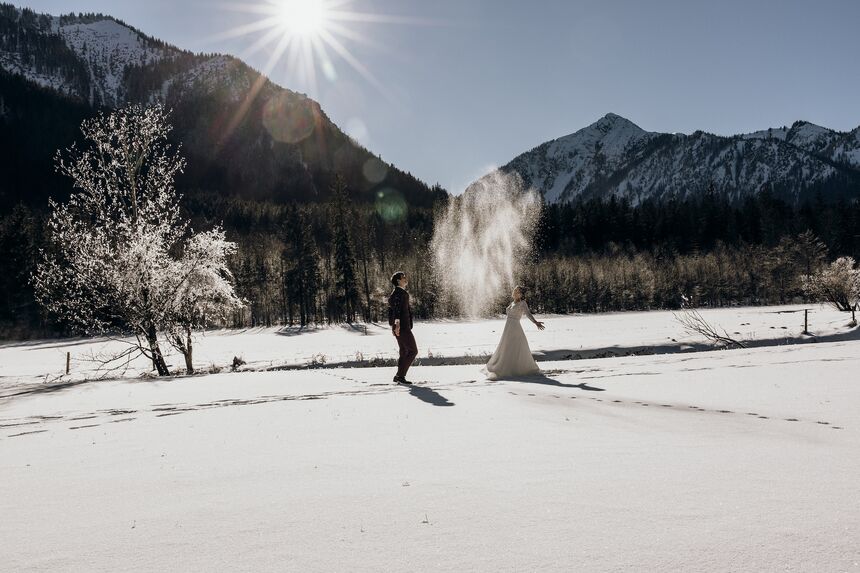 Winterhochzeit in den Bergen – Brautpaar spielt lachend im Schnee unter strahlender Sonne.