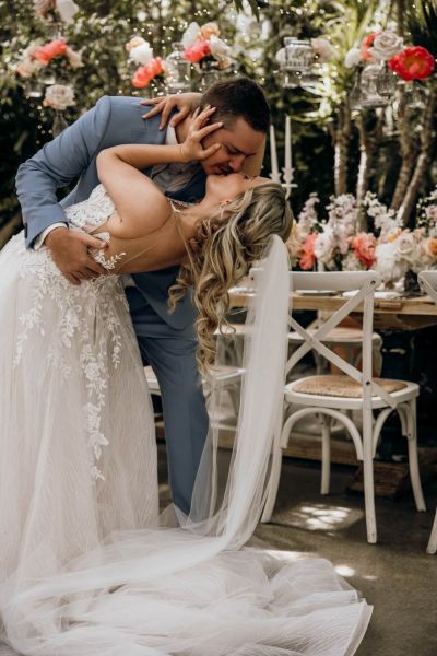 Bräutigam in hellblauem Anzug taucht Braut zum Kuss, romantische Szene mit Blumendekoration.