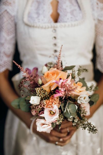 Braut in traditionellem Kleid hält einen bunten Hochzeitsstrauß mit Rosen und Eukalyptuszweigen.
