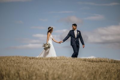 Braut und Bräutigam gehen Hand in Hand über ein Feld unter blauem Himmel – romantisches Hochzeitsfoto in natürlicher Umgebung.